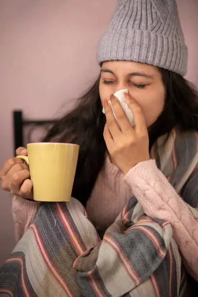 indoor image of a young woman getting sick with flu and cold and using a tissue paper to sneeze and blowing her nose while holding a coffee mug at home in winters. She is wearing warm clothes, sweater and cap.