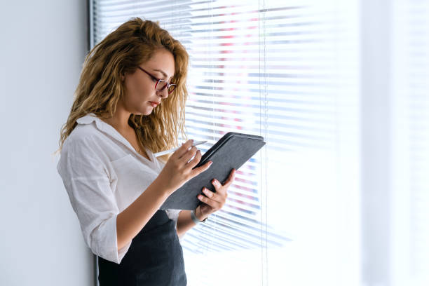 istock Young beautiful business woman is holding a mobile tablet on her hand, to check her work, while she is standing next to windows. 1340435787