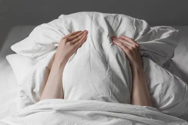 unrecognizable woman in bed covering face under pillow to hide from sunlight early in the morning