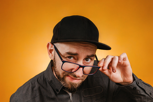 People and emotions. A man with a beard and glasses, wearing a black cap and shirt, stares through his glasses. Close up. Orange background.