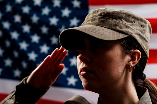 Memorial day. A portrait of female soldier in uniform salutes against the background of the American flag. Side view. The concept of the American national holidays and patriotism.