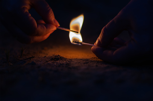 Two hands with burning matches in the dark. Man passes flame from lit wooden match to woman. Close-up, selective focus.