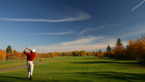 A fall golf scene. A man in red shirt hits a drive on a beautiful golf course in fall. Unrecognizable Caucasian senior male driving a golf ball on a rural valley course with beautiful golden colours. Man is showing good form and balance and proper golf technique. Back view. 