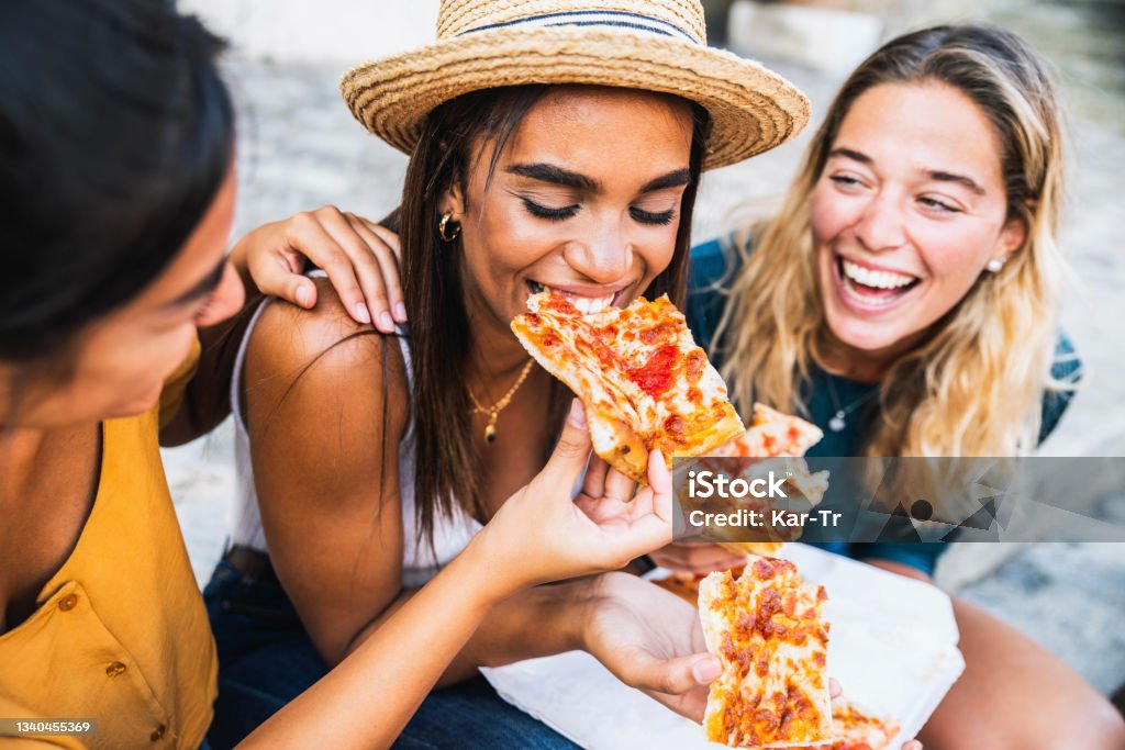 Three young female friends sitting outdoor and eating pizza - Happy women having fun enjoying a day out on city street - Happy lifestyle concept Pizza Stock Photo