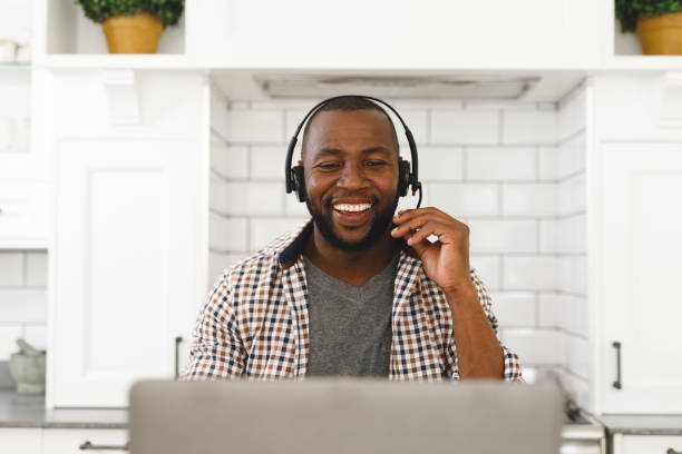istock Laughing african american man sitting in kitchen making video call using laptop and headset 1340459197