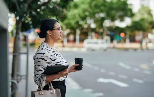 Young businesswoman with a coffee waiting for a taxi while standing on a sidewalk in the city in the morning