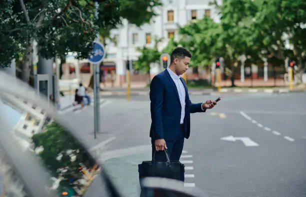 Young businessman checking an app on his phone while standing on a sidewalk in the city in the morning and waiting for a taxi