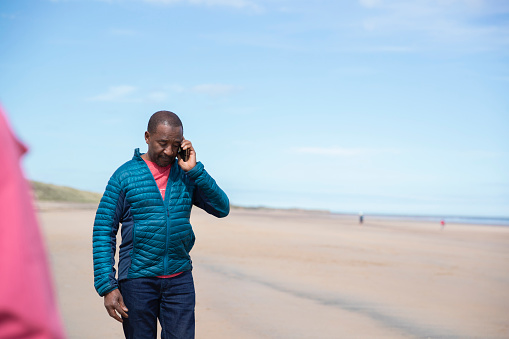 A senior black man wearing casual clothing on a beach on a sunny day in spring. He is standing and taking a phone call, he looks concerned.