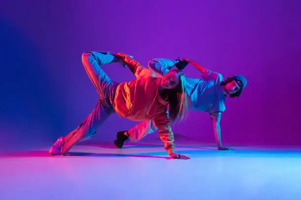 Stylish man and woman dancing hip-hop in casual sports youth clothes on gradient purple pink background at dance hall in neon light. Youth culture, hip-hop, movement, style and fashion, action.
