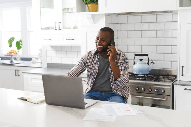 istock Smiling african american man sitting in kitchen working using laptop and smartphone 1340464275