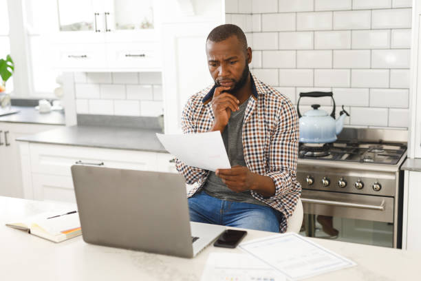 istock Serious african american man sitting in kitchen working looking at paperwork and using laptop 1340464276