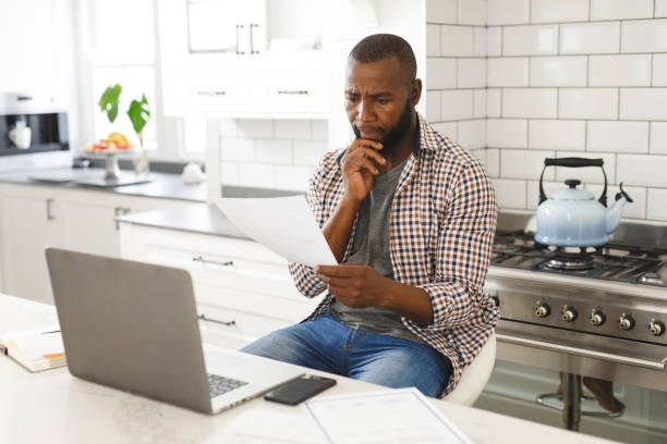 istock Serious african american man sitting in kitchen working looking at paperwork and using laptop 1340464336