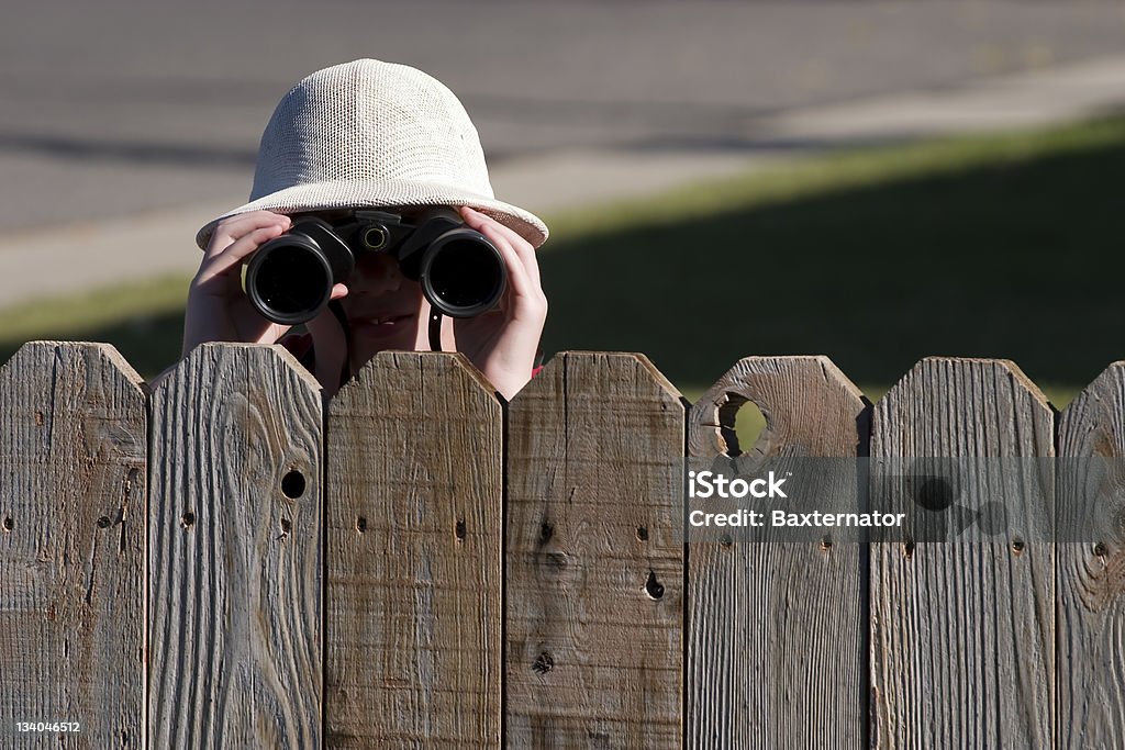 A neighbor dying over a fence wearing binoculars Young man wearing pith helmet looking with binoculars over fence. Fence Stock Photo