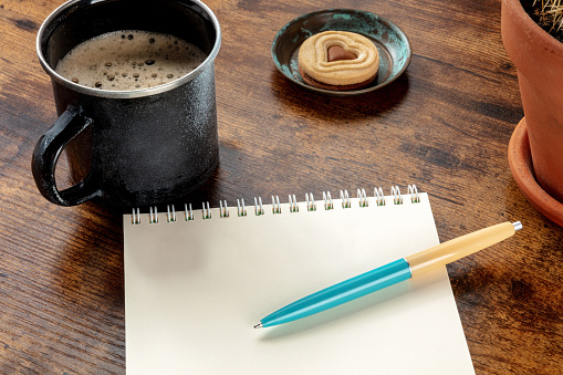 A blank notebook with a blue pen on a wooden desk, with a coffee mug and a chocolate cookie. Writing or planning
