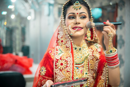 Beautiful happy traditional Indian bridal standing front of the mirror and getting ready for her wedding day. She is holding a makeup kit and makeup brush and doing her makeup herself.
