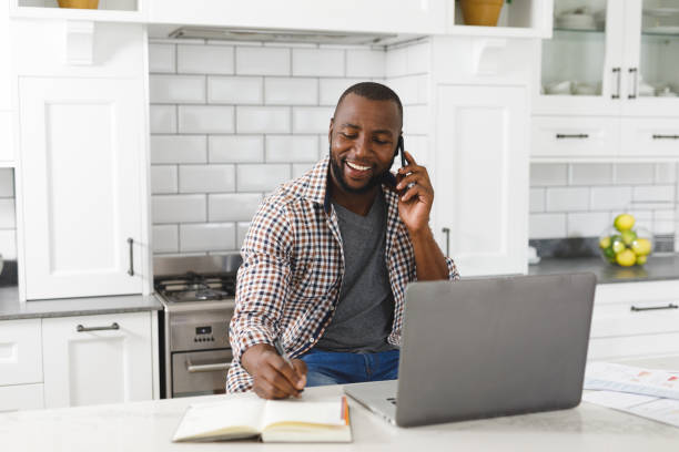 istock Smiling african american man sitting in kitchen working using laptop and smartphone 1340472319