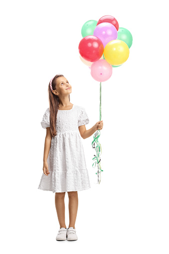 Full length portrait of a girl in a white dress holding a bunch of balloons and looking up isolated on white background