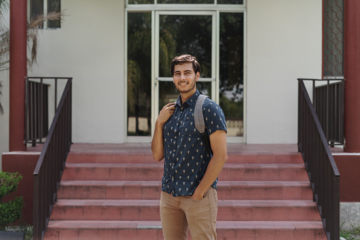 A happy latin male student standing outside a building and looking at the camera with a smile.