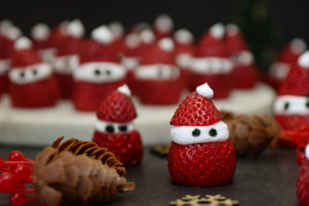 Stock photo showing close-up view of Christmas party themed food idea of strawberry and whipped cream Father Christmases. Strawberries are a popular fruit that is considered to be an especially healthy snack 'super food', since they are packed with beneficial antioxidants and fibre, being a good source of vitamin C. imagen en primer plano de la idea de comida de fiesta temática de navidad, santa claus de fresa frente al puesto de pasteles de mármol que contiene un lote de fresas de papá noel y crema batida, fondo negro de cono de pino, enfoque en primer plano