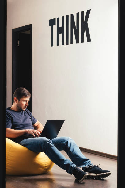 Handsome bearded man working on laptop and sitting on yellow bean bag soft armchair in modern office with the inscription think on white concrete wall