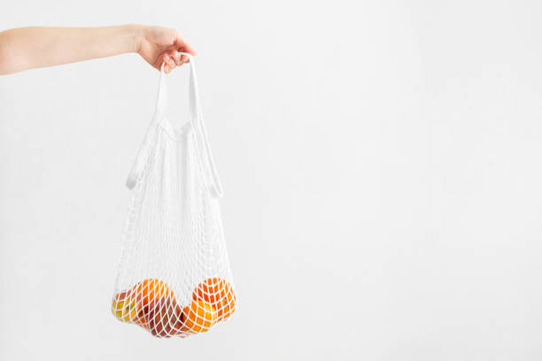 istock Hand holding white mesh bag with fruits on white background, copy space. Eco friendly, reusable shopping bag. Oranges, apples, garnet in cotton knitted string bag. Zero waste concept. 1340493453
