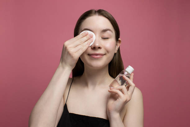 mujer joven sonriente con los ojos cerrados desmaquillando con almohadilla de algodón, sosteniendo una botella de agua micelar sobre fondo rosa. chica limpiando la cara. tratamiento y cosmetología. rutina de belleza, cuidado de la piel - human face water washing women fotografías e imágenes de stock