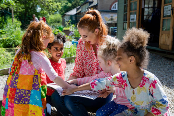 We Love This Story! A front view of a teacher and her excited little pupils sitting and doing storytime outdoors in the schoolyard on the benches on a wonderful summer's day in Hexham in the North East of England. elementary student pointing stock pictures, royalty-free photos & images
