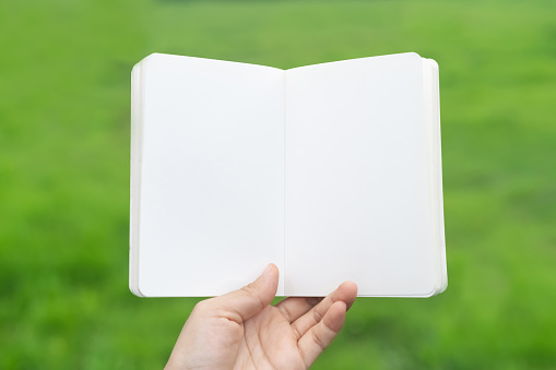 Copy space of man hand holding empty white notebook on green nature field abstract background.