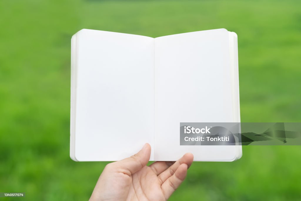 Copy space of man hand holding empty white notebook on green nature field abstract background. Note Pad Stock Photo
