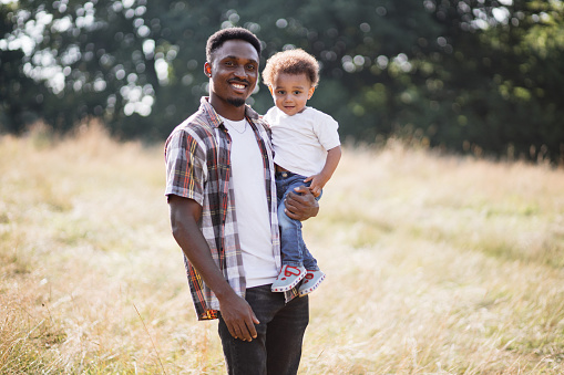 Front view of happy african american father holding his little son on hands while posing on summer field. Family connection and love concept.
