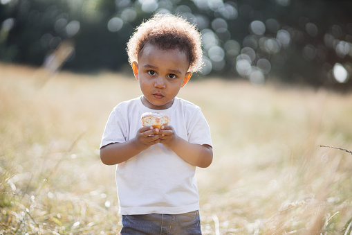 Portrait of pretty african american boy in white t-shirt and blue jeans holding sweet cupcake in hands while posing on summer nature. Sunlight in curly dark hair.