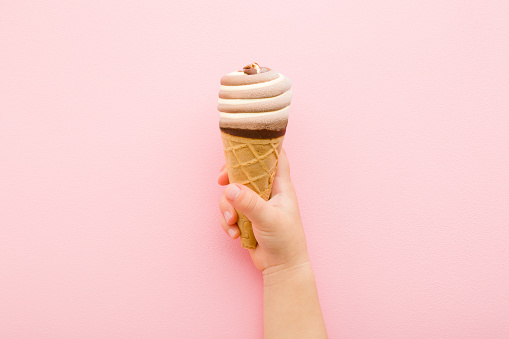 Baby hand holding waffle cone of striped black chocolate and white vanilla ice cream on light pink table background. Pastel color. Closeup. Beautiful cold sweet snack in summer. Top down view.