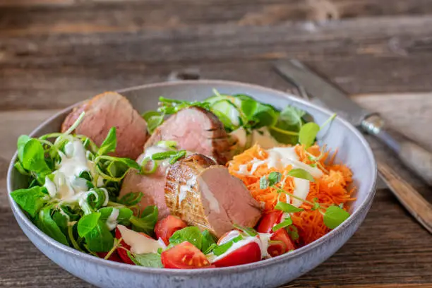Healthy low carb or ketogenic meal with slow cooked pork fillet and a mixed salad with yogurt dressing. Served isolated on a plate or bowl on rustic and wooden background
