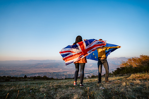 Two young pretty multi-ethnic women holding European Union and British flag on meadow. Brexit.