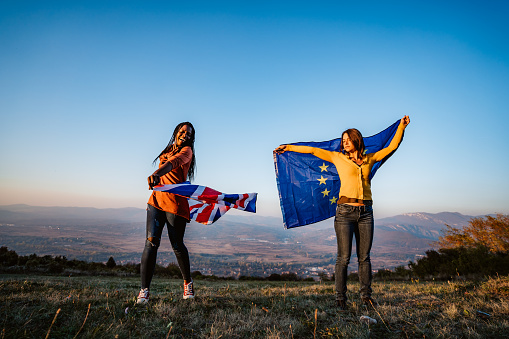 Two young pretty multi-ethnic women holding European Union and British flag on meadow. Brexit.