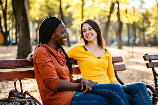Two female multi-ethnic friends talking on a park bench in autumn.
