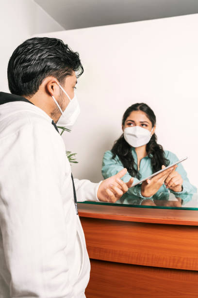 Receptionist with a mask attending to a man in a dental clinic Vertical photo of a young latina receptionist with a mask attending to a man in a dental clinic. office cubicle mask stock pictures, royalty-free photos & images