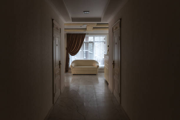 Stylish white empty sofa near large window in spacious living room view from semi-dark long corridor in contemporary house