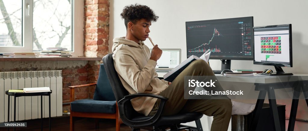 Banner view of ethnic male trader work online on computer Narrow web banner view of pensive African American millennial gen z male trader read book work online on PC at home. Serious young ethnic man financial expert analyze graph chart. Cryptocurrency. Stock Market and Exchange Stock Photo