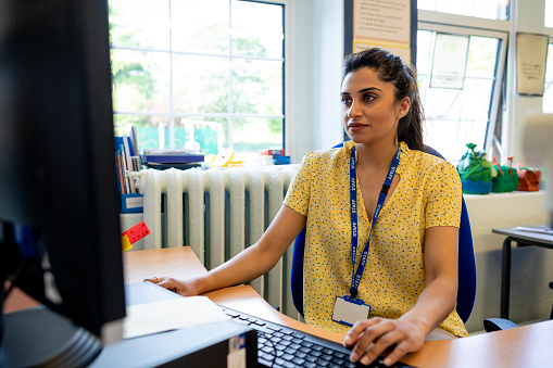 A close up front view of a young teacher sitting at her desk in her classroom. She is checking her lesson plans on her desktop PC. She is working in a school in Hexham in the North East of England.