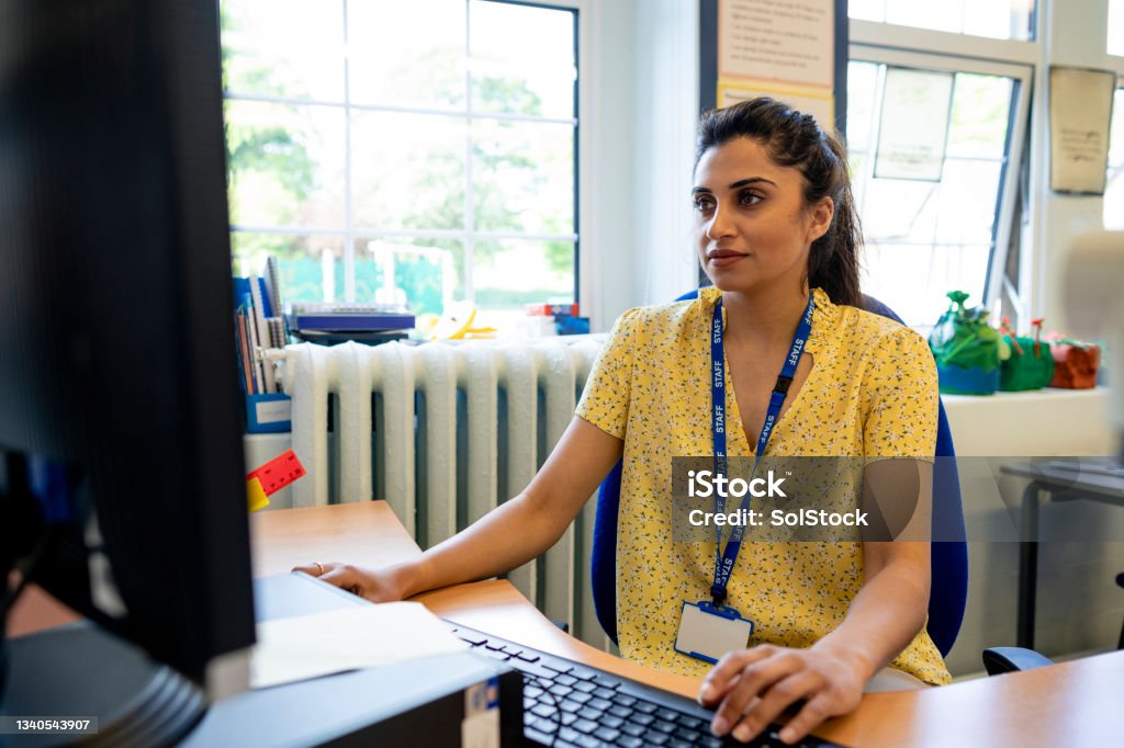 Teaching And Technology A close up front view of a young teacher sitting at her desk in her classroom. She is checking her lesson plans on her desktop PC. She is working in a school in Hexham in the North East of England. Teacher Stock Photo