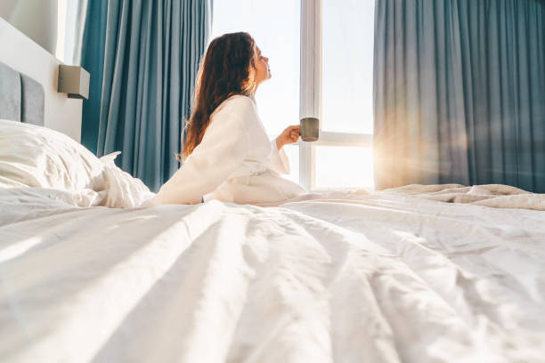 長い髪を持つかなりポジティブな女性は、日当たりの良い怠惰な朝に水を飲み、ベッドに座っています。 - ホテル ストックフォトと画像