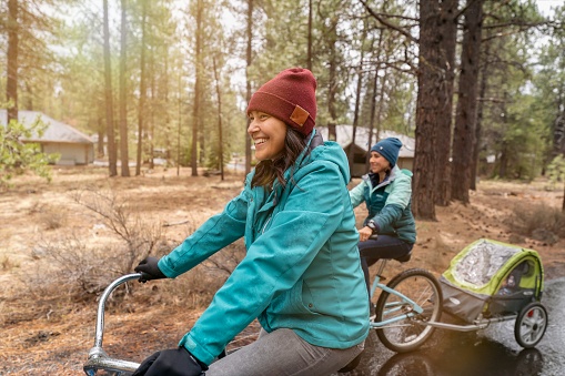 Two active mixed race Asian and Pacific Islander women enjoy a relaxing bike ride along a paved bike path in Oregon. The two women are sisters and one of the women is pulling her toddler in a carriage behind the bike. The smiling women are wearing jackets, gloves and winter hats because it is cold outside. The ground is wet from rain.