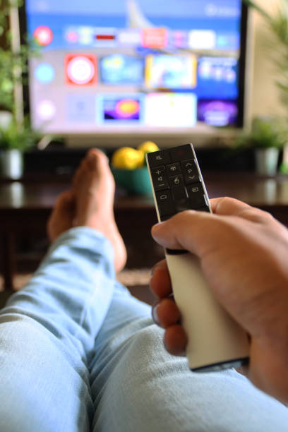 istock Image of unrecognisable person's hand holding a TV remote control, pointing at television screen, changing channels from rows of Smart Television Screen App Icons, legs stretched on coffee table 1340552032
