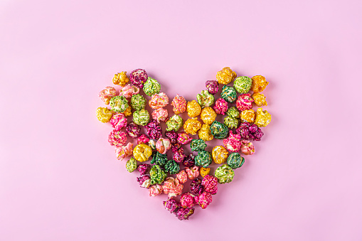 Love movies concept. Colorful rainbow caramel candy Popcorn scattered on pink background, heart shaped close up, copy space for text. Cinema snack concept