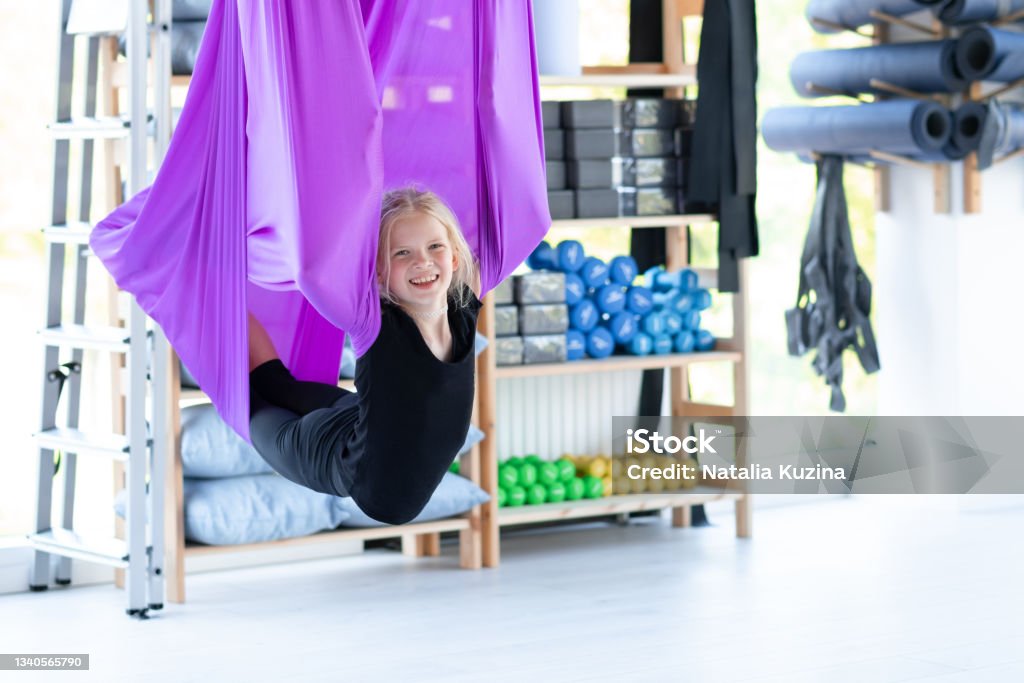 Portrait young smiling girl practice in aero stretching swing in purple hammock in fitness club. kids Aerial flying yoga exercises Portrait young smiling girl practice in aero stretching swing in purple hammock in fitness club. kids Aerial flying yoga exercises. Child Stock Photo
