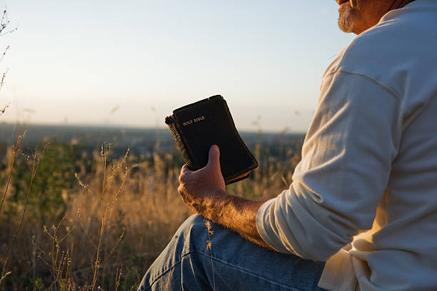 Alone, but not lonely. Man praying while holding a Holy Bible in an open field.  Bible stock pictures, royalty-free photos & images