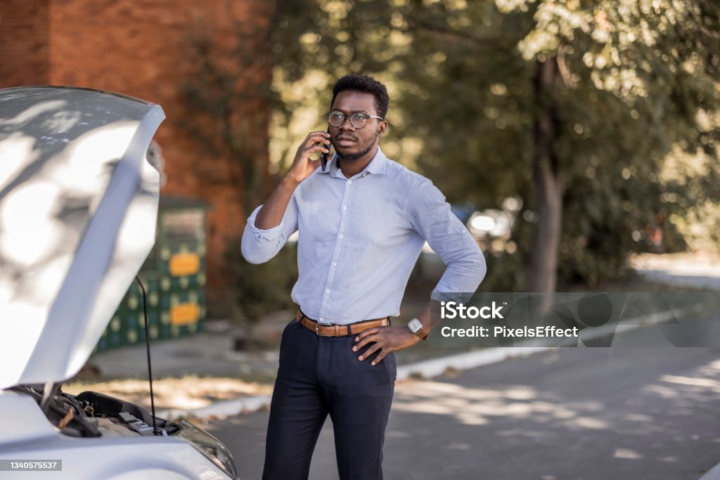 I need a little help Portrait of a young african man having trouble with his broken car calling for help on cell phone. Tow Truck Stock Photo