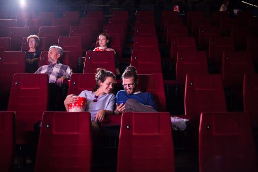 Young couple watching a comedy movie at the cinema and using a smartphone.