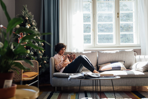 Smiling female sitting on couch next to the Christmas tree and looking at her smartphone